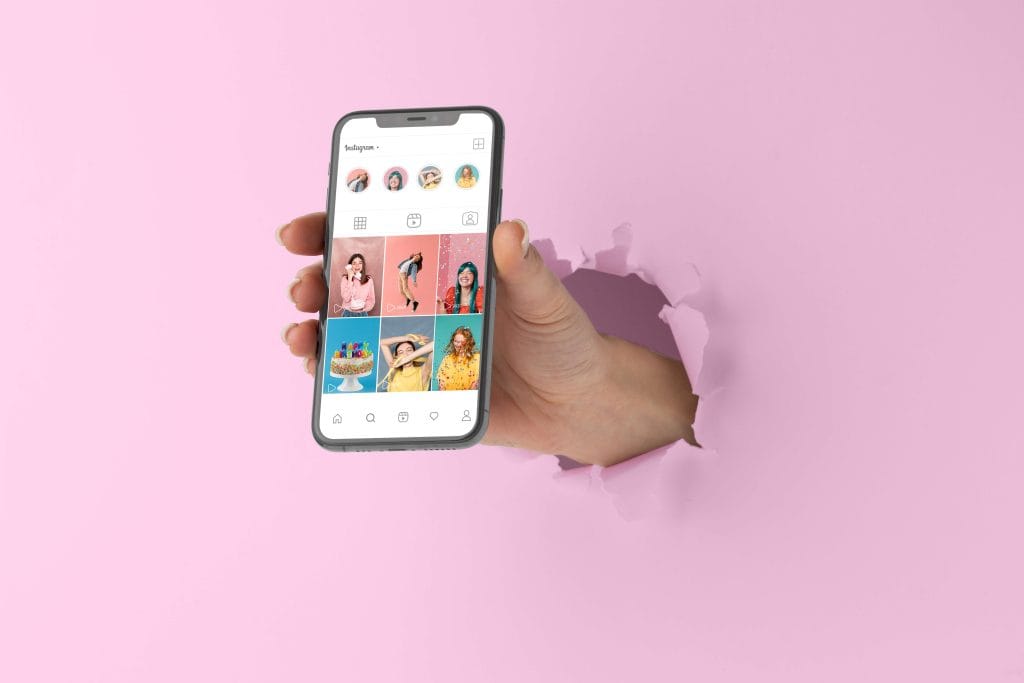 Fotografía de una mano saliendo por un agujero en una pared rosa, sosteniendo un teléfono móvil que muestra una cuenta de Instagram con fotos coloridas y creativas, en línea con servicios de diseño gráfico y redes sociales.