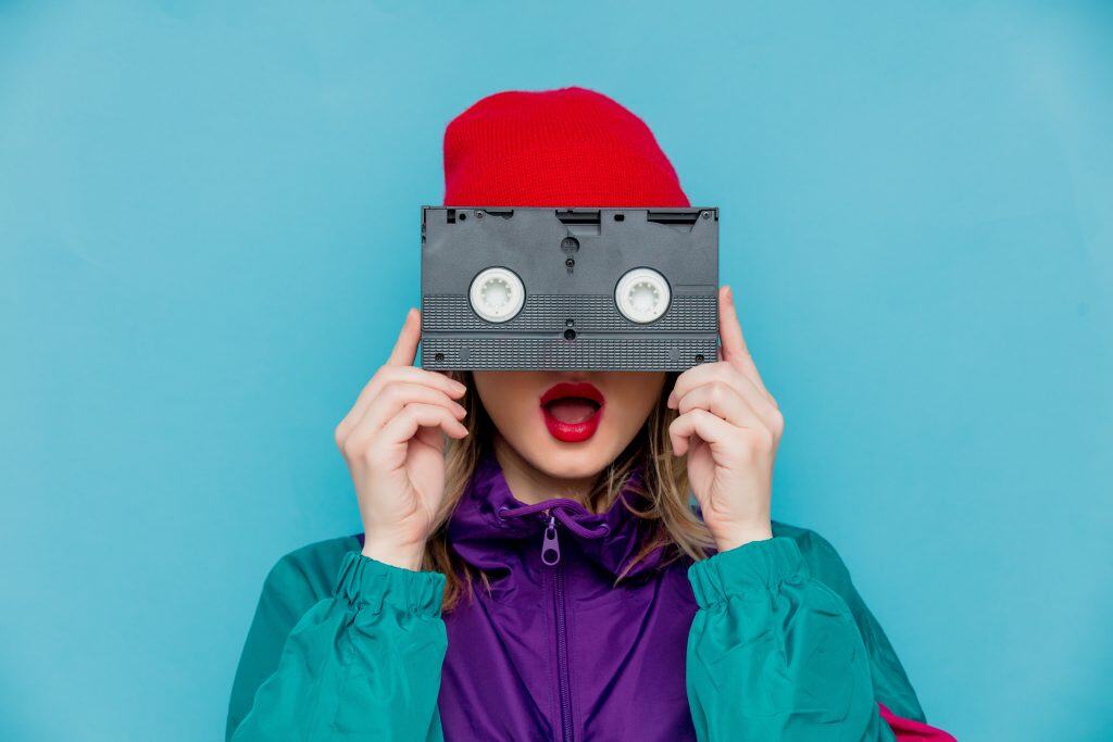Creativa mujer con gorro rojo y chaqueta multicolor sosteniendo una cinta de casete frente a la cara, fotografía moderna y artística, ideal para temas de cultura pop y música retro.